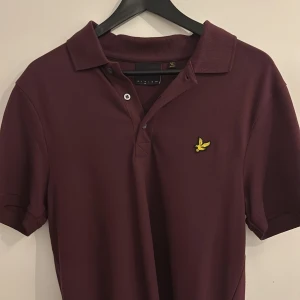 Vinröd pikétröja från Lyle & Scott - Snygg vinröd pikétröja från Lyle & Scott med klassisk krage och korta ärmar. Tröjan har tre knappar vid halsen och den ikoniska gula loggan broderad på bröstet. Perfekt för en stilren och avslappnad look.