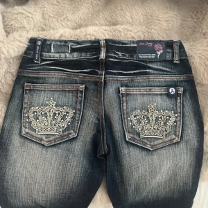 Crazy age jeans med kronbrodyr - Snygga blå jeans från Crazy Age med slitningar framtill och stora kronbroderier med pärldetaljer på bakfickorna. Jeansen har en klassisk femficksmodell, orangea sömmar och en rak passform. Perfekta för dig som vill sticka ut med coola detaljer.