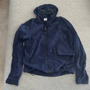 Marinblå Goggle Jacket från C.P. Company - Säljer en marinblå Goggle Jacket från C.P. Company med ikoniska glasögon i huvan. Jackan är tillverkad i lätt och slitstark nylon, har dragkedja framtill, knappdetaljer på fickan och justerbar huva. Perfekt för dig som gillar streetwear och snygga detaljer.