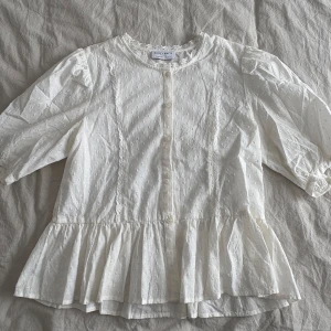 Vit blus med spets från Holly & Whyte - Superfin vit blus med spetsdetaljer vid halsen och knappar framtill. Blusen har trekvartsärmar, volang nertill och diskret strukturmönster i tyget. Köpte på Lindex, storlek S