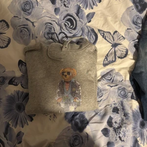 Grå hoodie Polo Bear från Ralph Lauren - Säljer en grå hoodie från Polo Ralph Lauren med den ikoniska Polo Bear broderad på bröstet. Tröjan har huva med snörning och är långärmad. Perfekt för dig som vill ha en chill och snygg streetstil. Nypris 2895kr, mitt pris 899