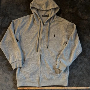 Grå polo Ralph Lauren zip hodie - Klassisk grå hoodie från Polo Ralp Laruen med dragkedja, huva med snörning och broderad logga på bröstet. Mjuk bomullsblandning, ribbade muddar och två fickor framtill. Perfekt för chill dagar eller när du vill ha en clean look.