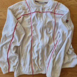 Vit ziptröja från FILA med röda detaljer - Vit långärmad ziptröja från FILA med röda linjer längs ärmar och bröst. Tröjan har dragkedja framtill, ståkrage med knapp och broderad FILA-logga på ärmen. Perfekt för en sportig och clean look. FILA TERRINDA PASSAR NÅN SOM ANVÄNDER XL. Finns i hässelby