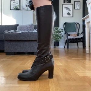 Svarta höga stövlar med klack, strl 37 - Säljer ett par svarta höga stövlar tillverkade i Italien. De har en rejäl klack, rund tå och dekorativa nitar samt spänne runt ankeln. Dragkedja på insidan för enkel på- och avtagning. Snygga till både jeans och kjol.