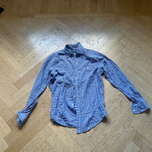 Blårutig skjorta från Ralph Lauren - Snygg blårutig skjorta från Ralph Lauren med klassisk krage och knappar framtill. Skjortan har lång ärm och gul broderad logga på bröstet. Tillverkad i mjuk bomull och har en custom fit-passform. Perfekt för dig som gillar stilrena och tidlösa plagg.