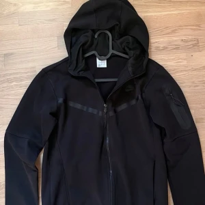 Svart Nike hoodiejacka med dragkedja - Svart hoodiejacka från Nike med hel dragkedja framtill och huva. Jackan har diskreta svarta detaljer över bröstet och en ficka med dragkedja på ena ärmen. Perfekt för en sportig och clean look.