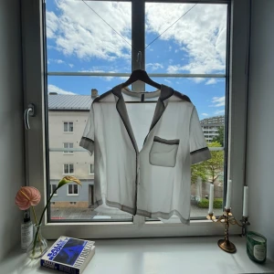 Vit kortärmad skjorta från Mango - Snygg vit skjorta från Mango med svarta kontrastkanter, kort ärm och bröstficka. Skjortan har en avslappnad passform och är tillverkad i mjukt material som känns svalt mot huden. Perfekt för en chill och stilren look.