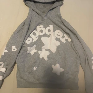 Grå hoodie med vita stjärnor från Sp5der - Grå hoodie från Sp5der med stora vita stjärnor och tryck på bröstet och ärmarna. Perfekt passform, huva och känguruficka på framsidan.