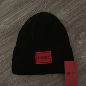 Svart mössa Hugo Boss med röd patch - Svart ribbstickad mössa från Hugo Boss med en tydlig röd patch framtill där det står HUGO i svart. Mössan är tillverkad i 100% akryl och har en klassisk uppvikt kant. Perfekt för dig som gillar stilrena accessoarer med tydlig logga.