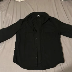 Svart overshirt från H&M i S - Svart overshirt från H&M med klassisk krage, två bröstfickor med lock och knäppning framtill. Skjortan är långärmad och har en rak, lite loose passform. Perfekt att slänga över en t-shirt för en chill look.