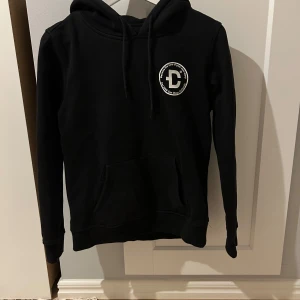 Svart hoodie med tryck från DirtCült - Svart hoodie från DirtCült med vit logga på bröstet. Klassisk modell med huva, dragsnören och magficka. Liten i storlek 