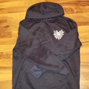 Svart hoodie med spindelnätsprint - Svart hoodie med stor vit spindelnätsprint på ryggen och mindre tryck på bröstet. Tröjan har huva med snörning och en klassisk känguruficka framtill. Perfekt för dig som gillar streetstyle och unika detaljer.