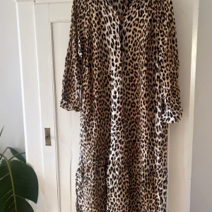 Leopardmönstrad långklänning - Säljer en snygg långklänning med leopardmönster och knappar framtill. Klänningen har trekvartsärm och en avslappnad passform som ger en cool vibe. Perfekt för dig som vill sticka ut med ett djuriskt print och samtidigt ha det bekvämt.