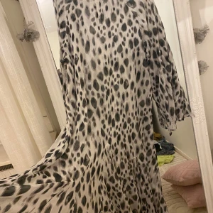 Långklänning med leopardmönster - Säljer en flowy långklänning med svart och grått leopardmönster på ljus botten. Klänningen har långa ärmar och är tillverkad i ett tunt, lätt material som ger ett snyggt fall. Perfekt för dig som vill sticka ut med ett djurmönster och gillar en luftig känsla.