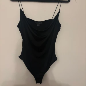 Svart bodysuit med tunna axelband Primark - Svart bodysuit från Primark med tunna axelband och snygg draperad urringning framtill. Tight passform som framhäver figuren och ger en clean look. Perfekt att styla med jeans eller kjol för en trendig outfit.