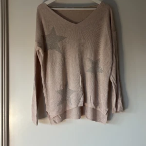 Beige stickad tröja med glittriga stjärnor - Rosa stickad tröja med v-ringning och glittriga silverstjärnor på framsidan och ena ärmen. Tröjan har en loose passform och långa ärmar, perfekt för dig som gillar en chill och trendig look. Materialet är mjukt och skönt mot huden.