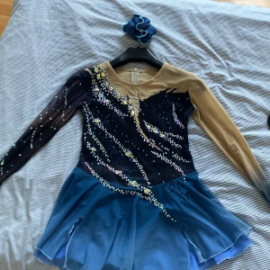 Blå glittrig konståkningsklänning - Supersnygg blå konståkningsklänning med glittriga stenar och meshdetaljer. Klänningen har långärmade ärmar i nude och svart mesh med kristaller. Har använt några enstaka gånger, inga defekter, kan få med extra stenar/kristaller. Priset kan diskuteras!