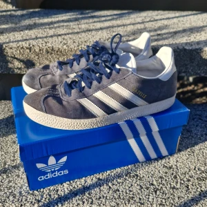 Adidas Gazelle blå sneakers mocka - Säljer ett par Adidas Gazelle sneakers i mörkblå mocka med vita detaljer och klassiska tre ränder på sidan. Skorna har rund tå, platt sula och snörning. Insidan är vit och de har en stilren retrovibe. Perfekt för dig som gillar ikoniska streetwear-skor. Om du har frågor är det bara att skriva till mig.