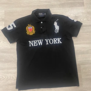 Svart pikétröja Ralph Lauren New York - Svart pikétröja från Ralph Lauren med broderat New York-emblem i guld, rött och blått på bröstet, stor vit polospelare och texten NEW YORK framtill. Nummer 5 i vitt på ärmen. Klassisk krage och knappar, tillverkad i bomull. Storlek S  den sitter perfekt på mig jag är 172cm. Den har inget defekt den är i jätte bra skick. Pris går att diskuteras 