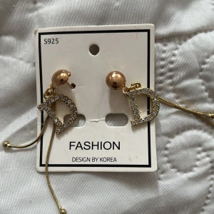 Guldiga örhängen med bokstaven D - Snygga örhängen i guldton med bokstaven D täckt av glittrande stenar. Hängande kedjor med små bollar ger extra bling och rörelse. Perfekta för dig som vill sticka ut med ett personligt smycke.