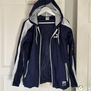 Blå vindjacka med huva från Puma - Snygg marinblå vindjacka från Puma med vita detaljer och dragkedjor. Jackan har huva med meshfoder, vita ränder längs ärmarna och två fickor med dragkedja. Perfekt för dig som gillar sportig stil och vill ha något lätt och funktionellt.