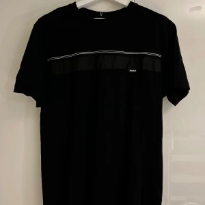 Svart t-shirt från Antony Morato - Svart t-shirt från Antony Morato med regular fit. T-shirten har en diskret vit rand över bröstet och ett litet Morato-tryck. Klassisk rund hals och korta ärmar. Materialet är mjuk bomull som känns skön mot huden.