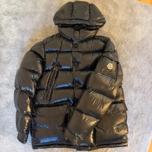 Svart Moncler pufferjacka med huva - Svart pufferjacka från Moncler med glansig finish, stor huva och feta fickor med knappar. Jackan har Moncler-logga på ärmen och dragkedja framtill. Perfekt för dig som vill ha en varm och stilren look under kalla dagar.