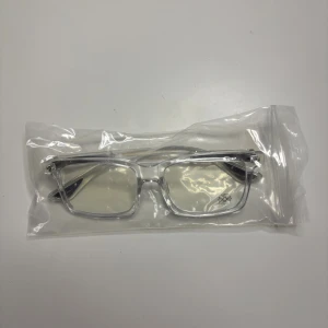 Transparenta rektangulära glasögon - Snygga glasögon med transparent rektangulär båge i plast och svagt tonade linser. På ena linsen finns en liten dekorativ blomma tryckt i svart. Perfekt för dig som vill ha en clean och modern look med en unik detalj.