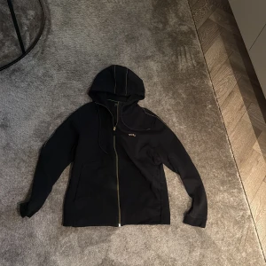 Svart hoodie från BOSS med dragkedja - Svart hoodie från BOSS i storlek S med guldfärgad dragkedja och huva. Snygga kontrastsömmar längs huvan och ärmarna samt diskret geometriskt mönster på ryggen. Mjuk bomullskänsla och klassisk passform, perfekt för chill dagar.