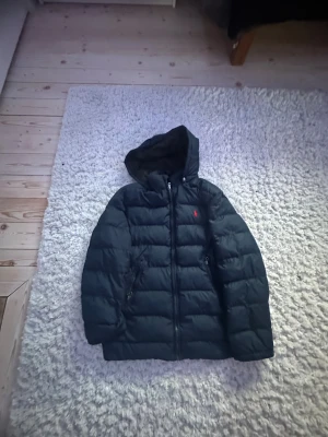Svart pufferjacka från Polo Ralph Lauren - Svart pufferjacka från Polo Ralph Lauren med huva och dragkedja framtill. Jackan har två sidofickor med dragkedja och den klassiska röda Polo-loggan broderad på bröstet. Materialet är syntet och jackan har quiltad design för extra värme.