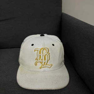 Vit keps Forever Phase 001 - Snygg vit keps med gul broderad logga på framsidan och coolt Forever Phase 001-märke på sidan. Justerbar snapback baktill och svarta detaljer på toppen och ventilationshålen. Perfekt för dig som vill sticka ut med en unik streetstyle-accessoar.