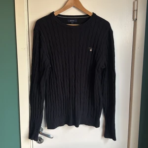 Svart kabelstickad tröja från GANT - Svart kabelstickad tröja från GANT i storlek M. Klassisk rund hals och diskret logga broderad på bröstet. Tröjan har långärmad passform och ribbade muddar vid ärmslut och nederkant. Perfekt för dig som gillar stilrena och tidlösa plagg.
