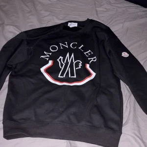 Svart Moncler sweatshirt med logga - Svart sweatshirt från Moncler med stor vit och röd logga på bröstet och Moncler-märke på ärmen. Tröjan har rund hals, lång ärm och är tillverkad i mjuk bomull. Perfekt för dig som gillar streetwear och vill ha något stilrent med tydlig branding.