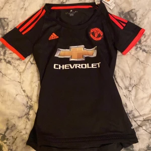 Manchester United svart matchtröja (dam tröja!) - Svart Manchester United fotbollströja från Adidas med röda detaljer och korta ärmar. Tröjan har klubbmärke på bröstet, Adidas-logga och Chevrolet som sponsor. Tillverkad i lätt och ventilerande ClimaCool-material, perfekt för träning eller match.