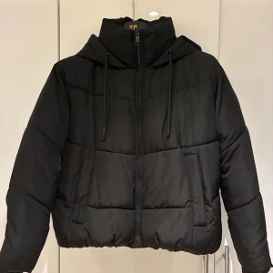 Svart pufferjacka från Zara XS - Svart pufferjacka från Zara i storlek XS. Jackan har huva med snörning, två sidofickor och dragkedja framtill. Den är croppad och har en stilren, boxig passform som ger en cool vibe. Perfekt för kalla dagar när du vill vara både varm och snygg.