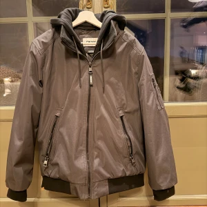 Svart vinterjacka från DKNY med huva - Snygg svart vinterjacka från DKNY med huva och flera dragkedjor. Jackan har ribbade muddar, fickor med dragkedja och en extra ficka på ärmen med DKNY-logga. Materialet är syntet och jackan har en modern, loose passform som passar till streetwear.