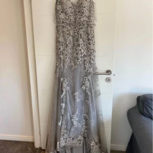 Grå långklänning med spetsdetaljer - Säljer en elegant grå långklänning med vacker spets och blommiga broderier. Klänningen har v-ringad urringning och är ärmlös, med ett skirt lager som ger en lyxig känsla. Perfekt för bal eller fest. Passformen är normal och materialet känns som polyester.och denna klängig är från L and s där ordinarie pris är 5000kr