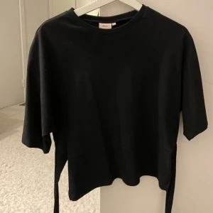 Svart topp med knyt från Nelly - Svart topp från Nelly med kort ärm och rund hals. Toppen har en cool omlott-detalj baktill som knyts i ryggen för en snygg siluett. Tillverkad i mjuk bomull som känns skön mot huden. Perfekt för dig som gillar stilrena och trendiga plagg.