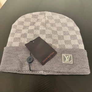 Grå rutig mössa - Snygg grå mössa med klassiskt rutigt mönster och uppvikt kant. logga framtill ger en lyxig touch. Mjuk och skön kvalitet, perfekt för kyliga dagar. Passar dig som vill ha en stilren och exklusiv look.