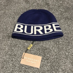 Blå Burberry mössa med logga - Snygg marinblå mössa från Burberry med stor vit logga runt hela kanten. Stickad i mjukt material som håller dig varm och ger en cool streetstil. Perfekt för dig som vill ha något trendigt och exklusivt på huvudet. London-vibe och klassisk design.