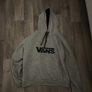 Grå hoodie från Vans med svart tryck - Säljer en grå hoodie från Vans med stort svart logotryck på bröstet. Tröjan har huva med svarta snören, känguruficka och mjukt material som känns skönt mot huden. Perfekt för chill dagar och streetwear-stil.