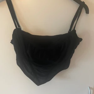 Svart croppad topp från SHEIN - Svart croppad topp från SHEIN med smala axelband och lätt rynkad volangdetalj framtill. Toppen har en enkel, stilren design och är tillverkad i ett tunt, mjukt material som känns skönt mot huden. Perfekt för att skapa en trendig look.