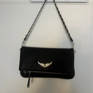 Svart axelväska med kedjerem - Snygg svart axelväska i skinnimitation med silverfärgad kedjerem och dekorativt vingemblem framtill. Väskan har dragkedja både fram och bak samt en stilren, avlång form. Köpt i Spanien förra sommaren för ca. 600kr. Använd 3/4 gånger så den är i väldigt fint skick 🤍