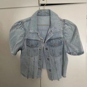 Ljusblå croppad jeansjacka med puffärm - Säljer en ljusblå croppad jeansjacka med korta puffärmar och rå kant nedtill. Jackan har klassisk krage, två bröstfickor med knappar och knappstängning framtill. Perfekt för dig som gillar unik denimstil och vill sticka ut.