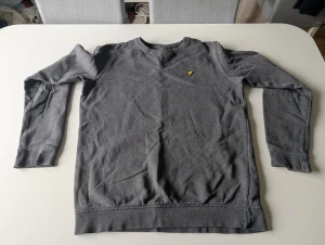 Grå långärmad tröja från Lyle & Scott - Säljer en stilren grå långärmad tröja från Lyle & Scott med klassisk rund halsringning och den ikoniska gula örnloggan broderad på bröstet. Tröjan är i mjuk bomull och passar perfekt till jeans eller chinos.