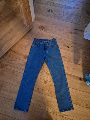 Levi's 501 blå jeans W25 L28 - Klassiska Levi's 501 jeans i blå tvätt med raka ben och fem fickor. Modellen har medel midja och är tillverkad i slitstark bomull. Perfekt passform och tidlös look, med den ikoniska läderpatchen bak i midjan.