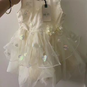 Champagnefärgad hjärtklänning Hi Fi - Superfin champagnefärgad kortklänning från Hi Fi Vibrama med glittriga hjärtan och två dekorativa blommor i midjan. Klänningen är ärmlös och har flera lager tyll som ger en fluffig och festlig look. Perfekt för speciella tillfällen!