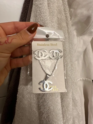 Set med halsband och örhängen - Säljer ett matchande set med halsband och örhängen i rostfritt stål, båda med den ikoniska CC-loggan. Smyckena är silverfärgade och har en stilren, modern look som passar perfekt till din outfit.