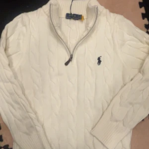 Vit stickad tröja Polo Ralph Lauren - Kabelstickad vit tröja från Polo Ralph Lauren med halv dragkedja och broderad marinblå logga på bröstet. Tröjan har ribbade muddar och hög krage, perfekt för lager-på-lager. Klassisk och stilren design som funkar året runt.