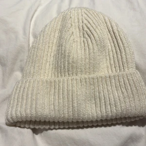 Vit ribbad stickad mössa från H&M - Snygg vit mössa från H&M med ribbad stickning och uppvikt kant. Perfekt för kalla dagar och enkel att matcha med din vinterstil. Mjuk och skön, tillverkad i stickat material som håller dig varm. oanvänd